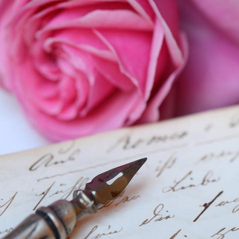 Federhalter mit Schreibfeder auf Buch mit alter Handschrift: Liebesbriefe sind Zeugnisse ihrer Zeit und Gesellschaft. Hieß es früher: „Ich liebe Dich“, werden heute Emojis mit Herzchen und Küsschen gesendet.