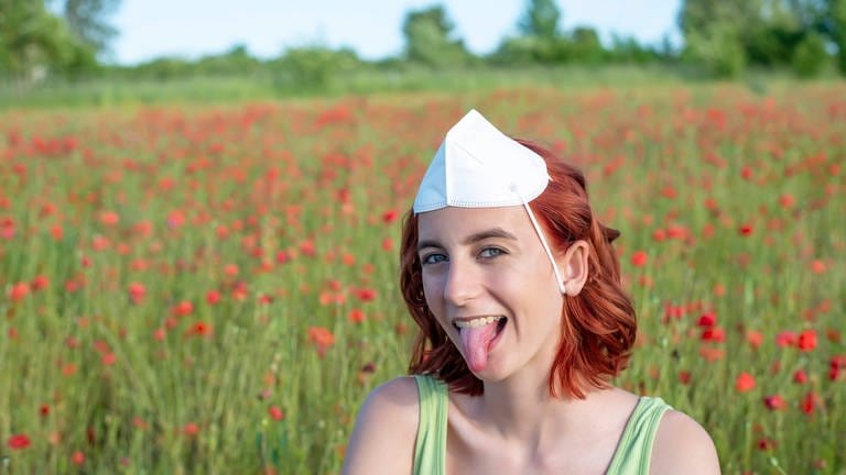 Mädchen mit Maske auf dem Kopf streckt in einem Mohnfeld fröhlich die Zunge heraus: 