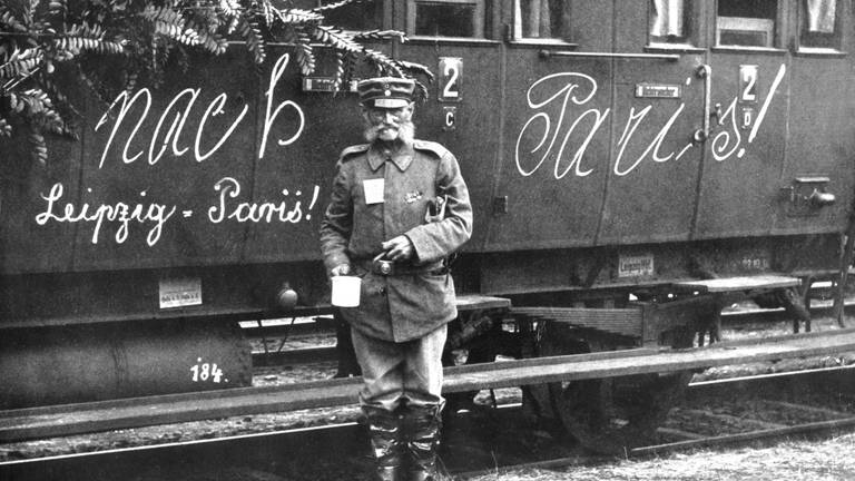 Soldat im Ersten Weltkrieg vor einem Zug mit der Aufschrift "Nach Paris – Leipzig  Paris!"