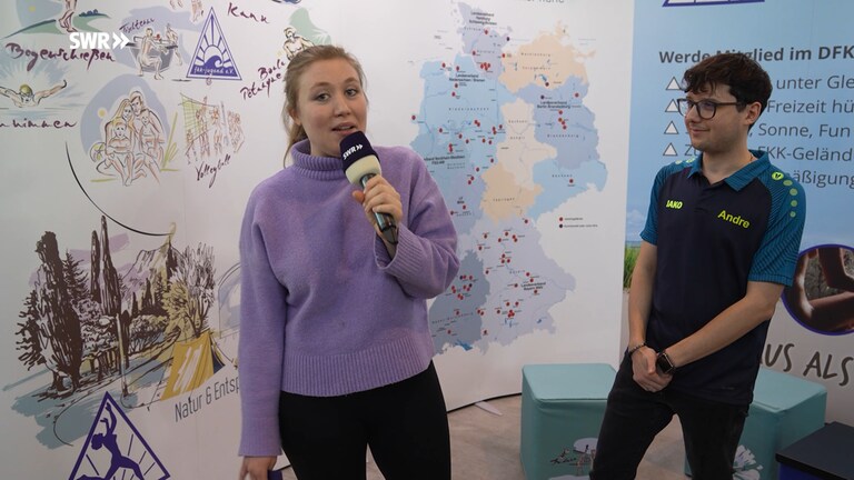 Eine Reporterin spricht in ein Mikrofon. Neben ihr steht ein Mann, der sie ansieht. Im Hintergrund ist eine Landkarte zu sehen, die beiden stehen am Stand des FKK-Vereins. (Foto: SWR)