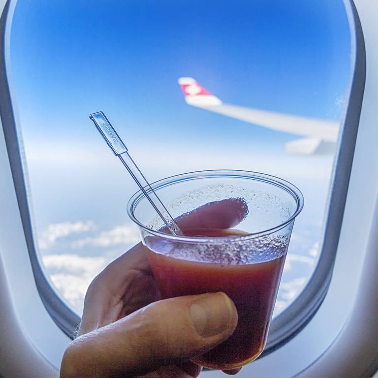 Eine Hand hält einen Becher mit Tomatensaft vor einem Fenster mit Blick aus einem Flieger: Der Geschmackssinn verändert sich im Flugzeug. Das wird beim Tomatensaft besonders deutlich.