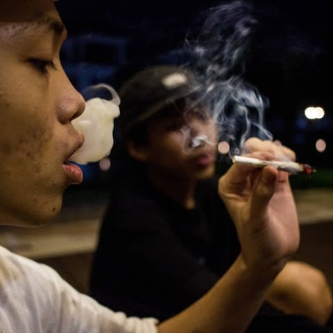 Jugendlicher atmet Rauch aus und hält einen glühenden Joint.
