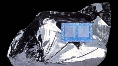 Auf einem unförmigen Stück metallisch glänzendem Silizium liegt ein bläulicher Mikrochip. (Foto: IMAGO, IMAGO / agefotostock)