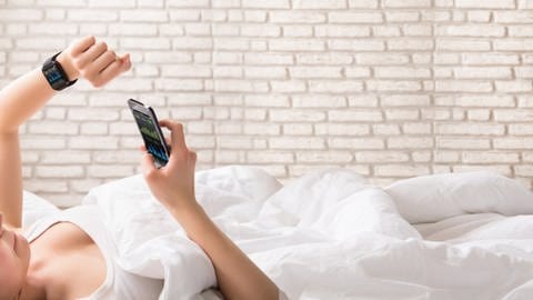 Frau liegt auf Bett und schaut auf ihre Smartwatch und ihr Smartphone.