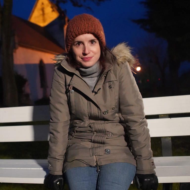 Junge Frau in Winterkleidung auf weißer Bank vor Kirche bei Nacht mit Blick in die Kamera (Foto: SWR)