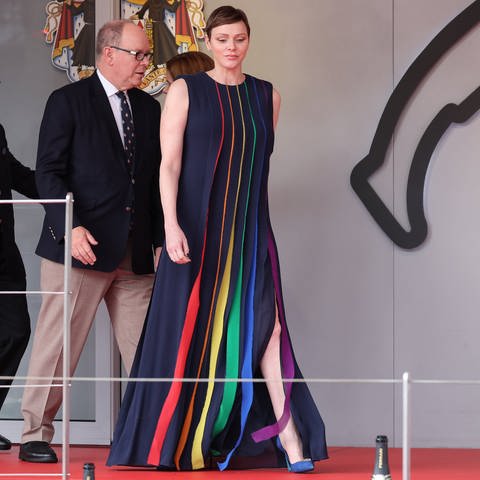 Charlène von Monaco trägt ein langes dunkelblaues Kleid, darauf sind Streifen in Regenbogenfarben zu sehen. Sie schreitet voran auf der Formel 1 Bühne in Monaco, wo das Rennen stattfand. Direkt hinter ihr läuft ihr Mann Fürst Albert von Monaco in einem schicken Anzug.