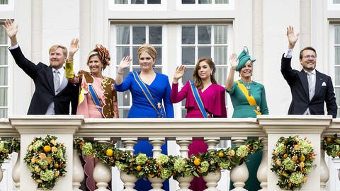 Die niederländische Königsfamilie steht auf einem mit Blumen geschmücktem Balkon. Von links nach rechts winken in schicken Kleidern: König Willem-Alexander, Königin Maxima, Kronprinzessin Amalia, Prinzessin Alexia, Prinzessin Laurentien und Prinz Constantijn. 