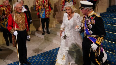 Mann im Anzug mit Orden und Hut führt Frau im langen weißen gold bestickten Kleid mit Krone an der Hand auf blauem Teppich entlang