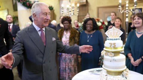 König Charles feiert 75. Geburtstag schon vor - mit einer großen Torte: Der britische Monarch steht mit ausgebreiteten Armen vor einer dreistöckigen Geburtstagstorte mit der Zahl 75 darauf. Im Hintergrund sind unscharf mehrere Menschen zu sehen.