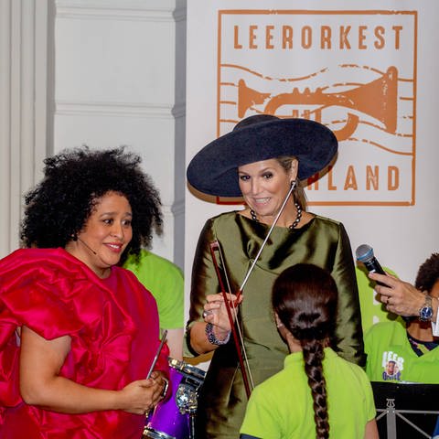 Königin Maxima und die Mezzosopranistin Tania Kross besuchen das Symposium des Lernorchesters im Concertgebouw in Amsterdam. Sie steht mit einem Tacktstock vor einem Mädchen und lächelt sie in einem grünen Dress mit großem Hut an.