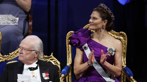 Frau in langem lila Ballkleid mit großer Blume auf der Schulter mit Krone applaudiert, links vor ihr sitzt ein Mann mit Brille und grauem Haar - Prinzessin Victoria von Schweden und König Carl Gustaf