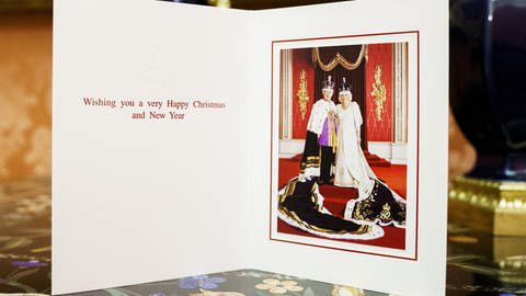 Ein Foto von König Charles und Königin Camilla in einer Klappkarte, sie stehen in königlicher Robe mit Kronen im Palast auf einer roten Treppe