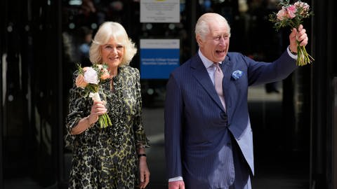 König Charles III. und seine Frau Königin Camilla besuchen gut gelaunt eine Krebszentrum. Beide tragen kleine Sträuße in der Hand.