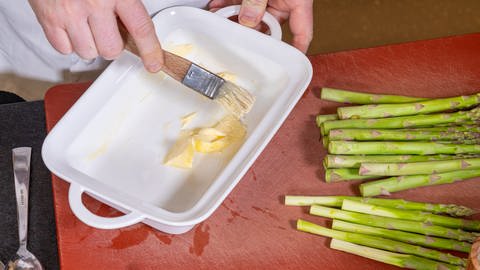 Eine weiße Auflaufform steht auf einem roten Brett und wird mit einer Hand mit Butter eingepinselt, daneben liegen grüne Spargelstangen