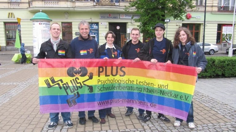 Menschen halten gemeinsam einen bunten Banner in Regenbogenfarben hoch. Die Aufschrift: PLUS, psychologische Schwulen- und Lesbenberatung Rhein-Neckar