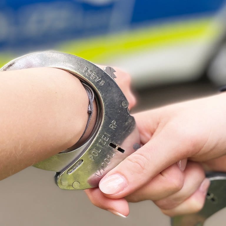 Eine Polizistin legt einem Mann Handschellen an. Im Hintergrund ist ein Polizeiauto zu sehen.