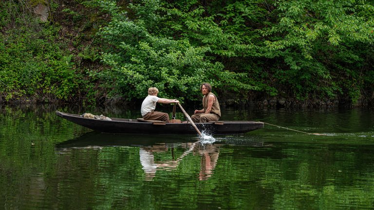 Thor und Ola auf einem Holzboot in einem Gewässer. Thor (Jenö Stillmark) rudert, Ola (Maximilian Klas) angelt Fische.