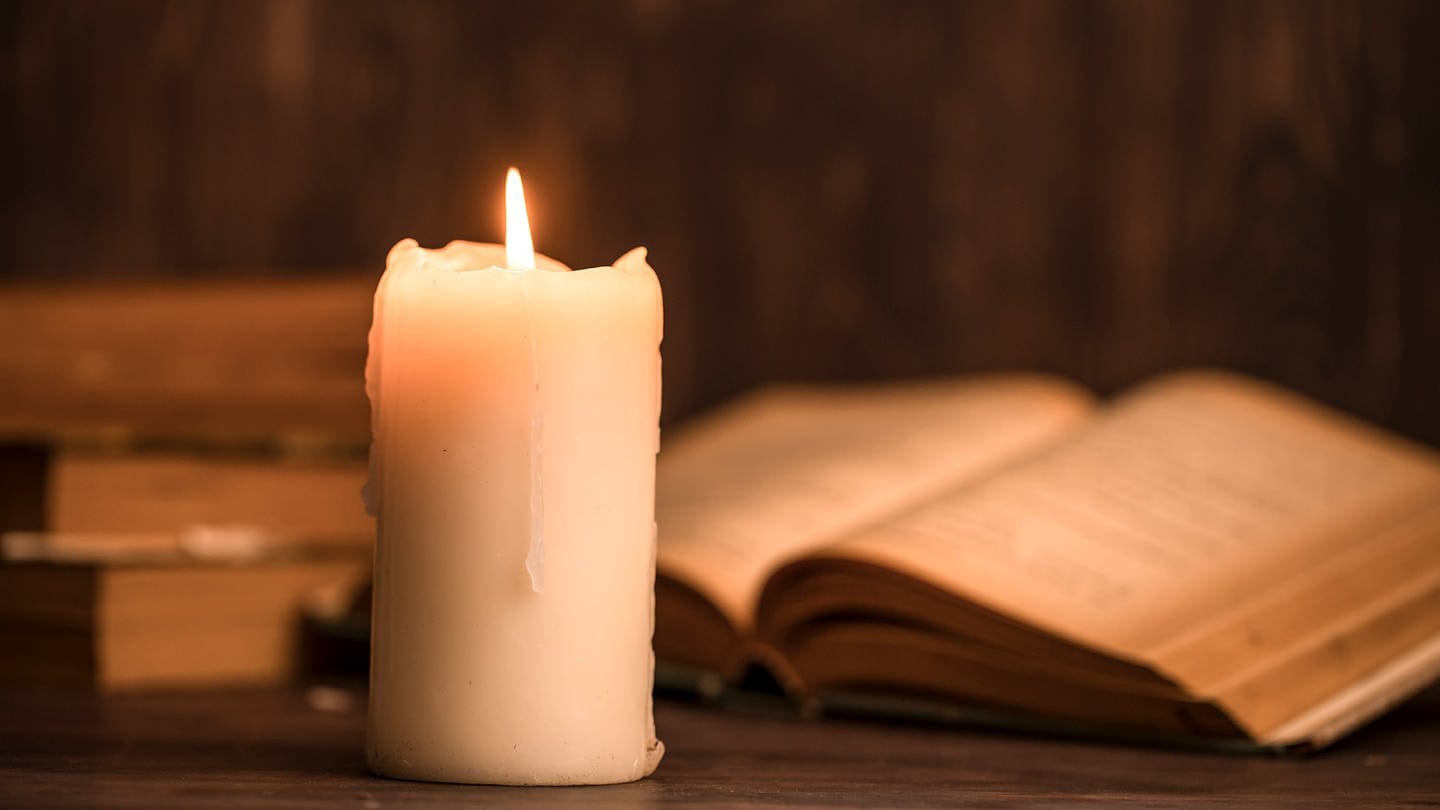 Eine brennende Kerze steht vor einem aufgeklappten Buch (Foto: Getty Images, Photobylove)