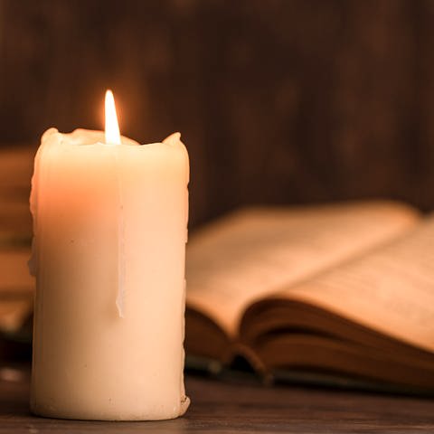 Eine brennende Kerze steht vor einem aufgeklappten Buch (Foto: Getty Images, Photobylove)