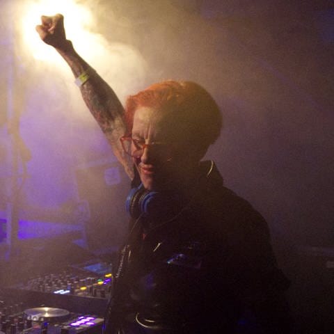 DJ Elly steht auf einer Bühne hinter dem DJ Mischpult. Sie streckt in Feierlaune eine Faust in die Luft.  (Foto: Cora Schäfer)
