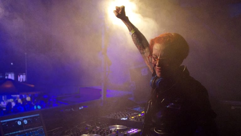 DJ Elly steht auf einer Bühne hinter dem DJ Mischpult. Sie streckt in Feierlaune eine Faust in die Luft.  (Foto: Cora Schäfer)