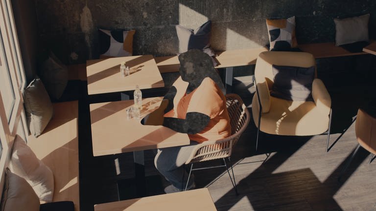 Eine Frau sitzt alleine im Cafe. Sie erzählt über ihre Adoption und ist anonym dargestellt. Ihr Gesicht und ihre Hände sind unkenntlich gemacht.