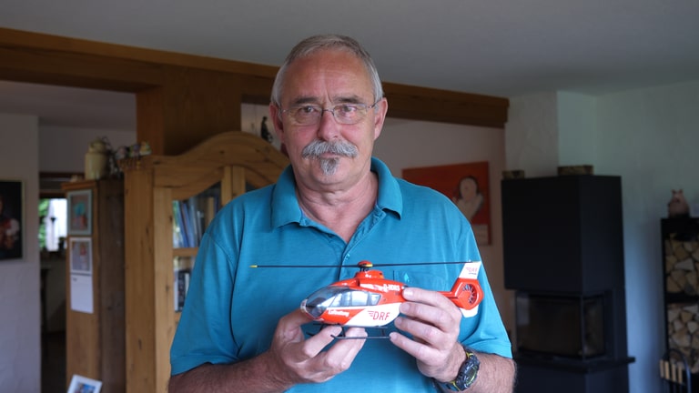 Hubschrauberpilot Andreas in seinem Wohnzimmer, er hält ein Modell eines DRF-Hubschraubers in den Händen und schaut direkt in die Kamera