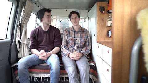 Zwei junge Männer sitzen in einem Bus nebeneinadner und unterhalten sich (Foto: SWR)