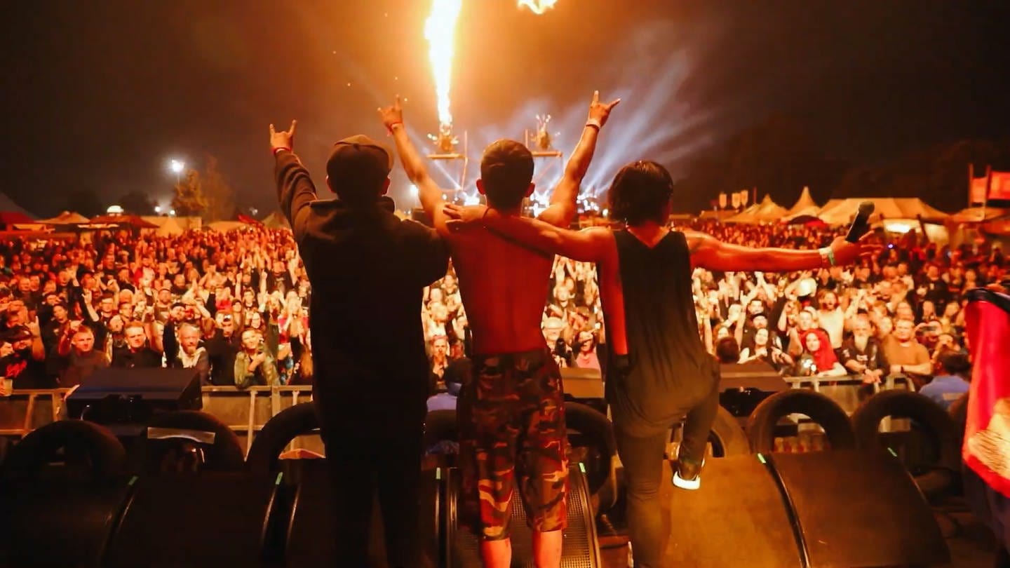 Drei Musiker von hinten, stehen auf der Bühne in Wacken, halten sich in den Armen, Publikum jubelt (Foto: SWR)