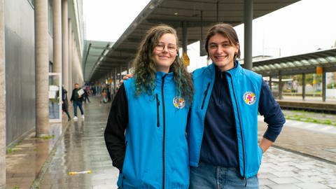 Sinja und Ellen machen ihr Freiwilliges Soziales Jahr bei der Bahnhofsmission in Freiburg. Sie tragen die Arbeitskleidung der Bahnhofsmission und lachen in die Kamera.  (Foto: SWR)
