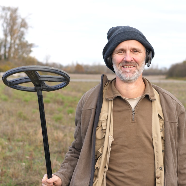 Gerhard ist Hobby Archäologe und hält einen Metalldetektor in der Hand und lächelt in die Kamera.