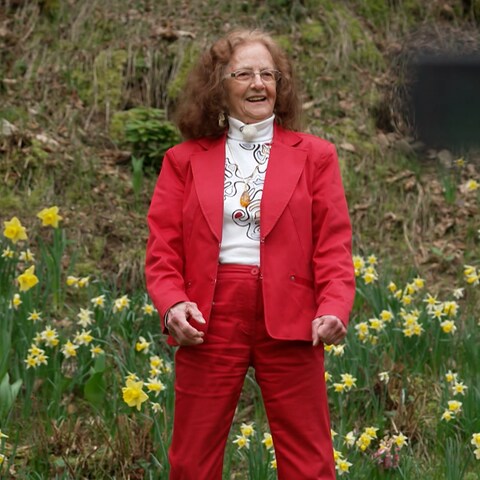 Thea rockt - ältere Frau in rotem Kostüm vor blühender Hecke (Foto: SWR)