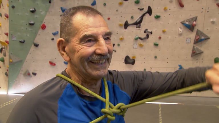 Der 89-jährige Franz steht in Sportkleidung vor einer Kletterwand in einer Kletterhalle und lächelt. (Foto: SWR)