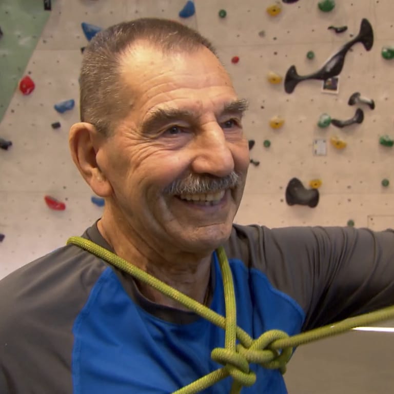 Der 89-jährige Franz steht in Sportkleidung vor einer Kletterwand in einer Kletterhalle und lächelt. (Foto: SWR)