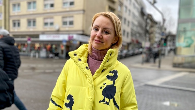 Kateryna aus der Ukraine steht in Innenstadt von Pforzheim. Sie trägt einen schrillen gelben Mantel und schaut in die Kamera. 
