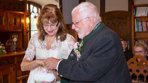 Edith und Harald steigen bei ihrer Hochzeit durch ein großes Herz. Sie trägt ein weißes Dirndl und er eine braune Lederhose. 