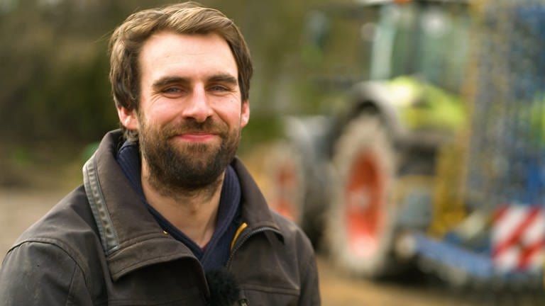 Junger Mann mit langem Bart, brauner Jacke vor einem Traktor (Foto: SWR)