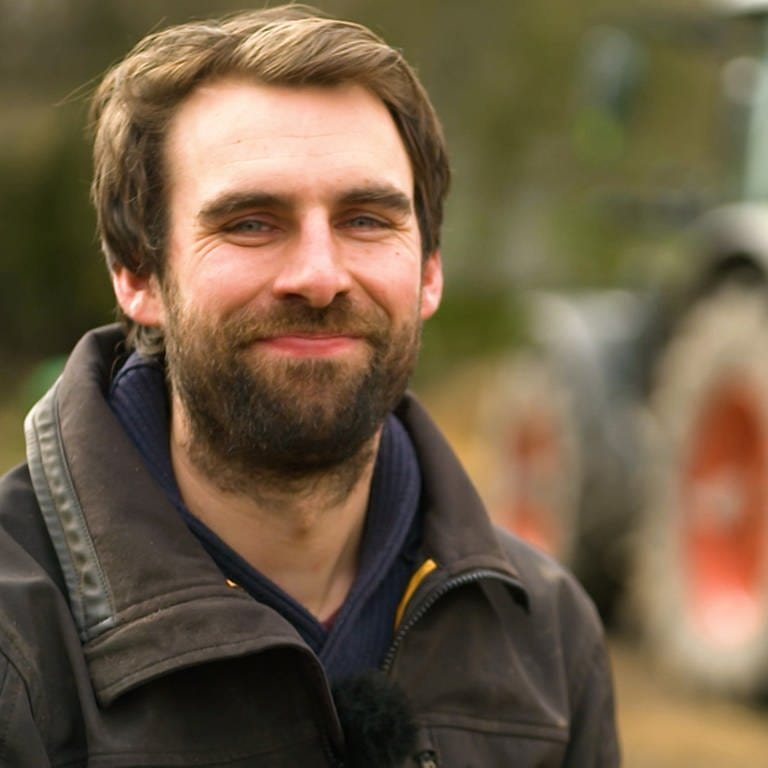 Junger Mann mit langem Bart, brauner Jacke vor einem Traktor