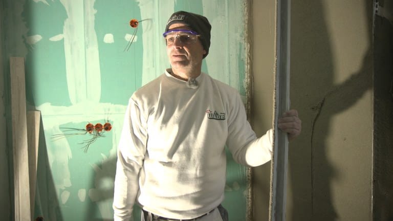Stuckateur Manfred ist auf einer Baustelle. Er trägts seine Arbeitskleidung und eine Schutzbrille. Im Hintergrund stehen Kabel aus den Wänden.  (Foto: SWR)
