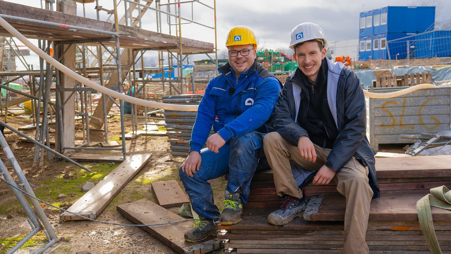 Zwei junge Männer sitzen in Arbeitskleidung auf einer Baustelle und lächeln in die Kamera. (Foto: SWR)