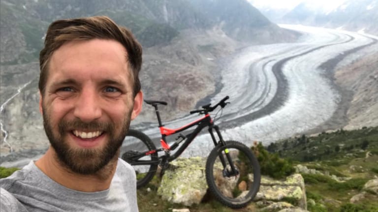 Junger Mann mit braunen Haaren lächelt in die Kamera, im Hintergrund Berge und ein Fahrrad (Foto: SWR)