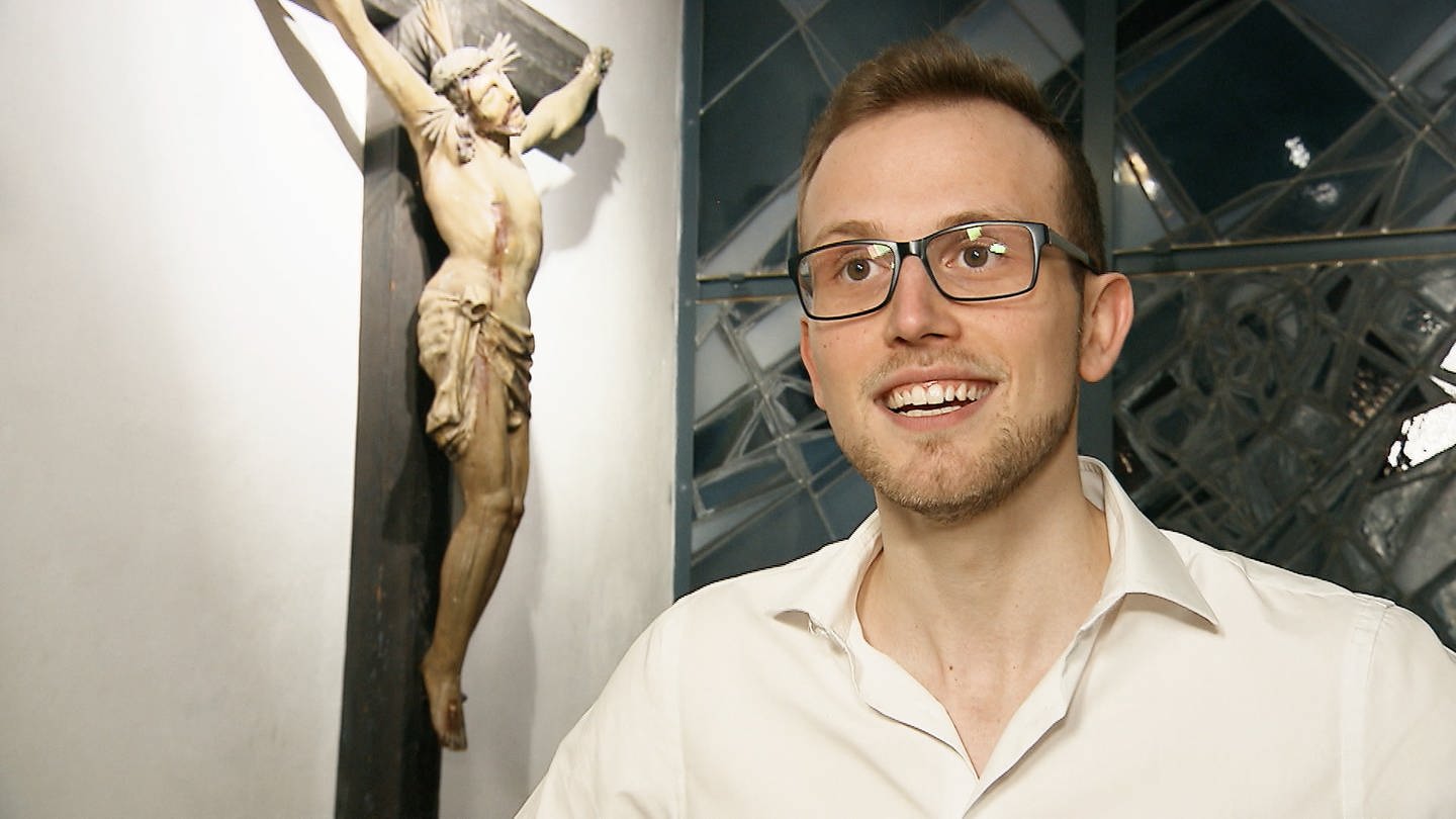 Junger Mann mit Brille steht in einer Kirche vor einem Holzkreuz.
