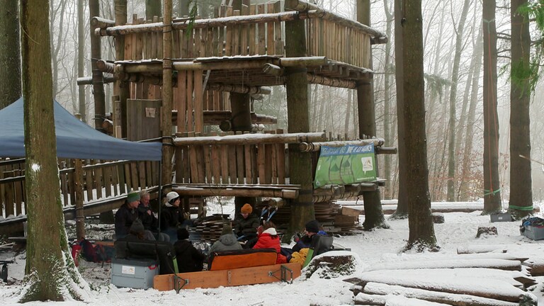 Im Vordergrund eine Gruppe junger Menschen, dick eingepackt, an einem Lagerfeuer. Im Hintergrund ein Wald mit einem mehrstöckigen Baumhaus.  (Foto: SWR)