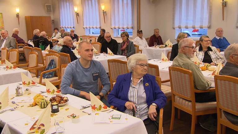 Ein nobler Essenssaal, im Vordergrund sitzt ein junger Mann neben einer älteren Dame an einem Tisch. (Foto: SWR)