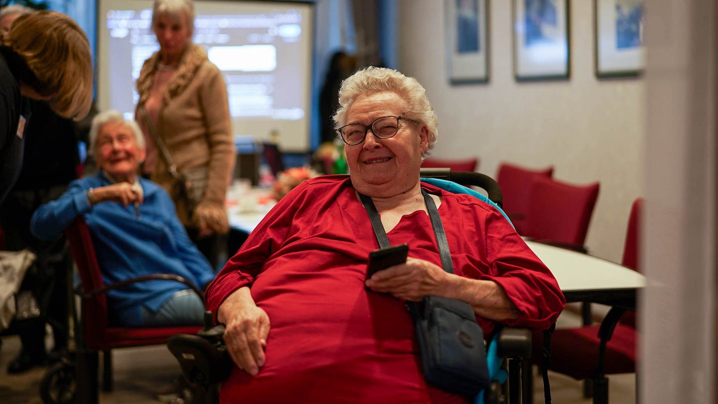 Ursula, eine ältere Frau, die im Rollstuhl sitzt. In ihrer Hand zeigt sie ihr Handy. (Foto: SWR)