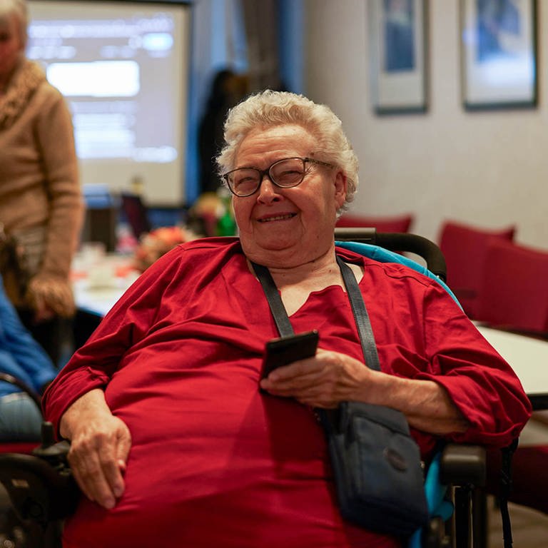Ursula, eine ältere Frau, die im Rollstuhl sitzt. In ihrer Hand zeigt sie ihr Handy. 