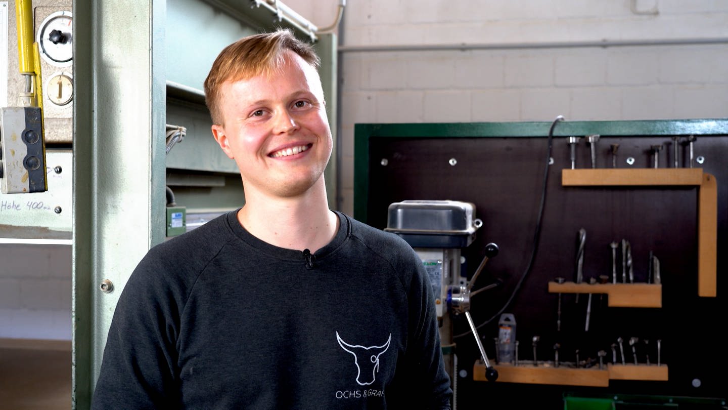 Ein junger, blonder Mann steht in einer Werkstatt und lächelt in die Kamera (Foto: SWR)