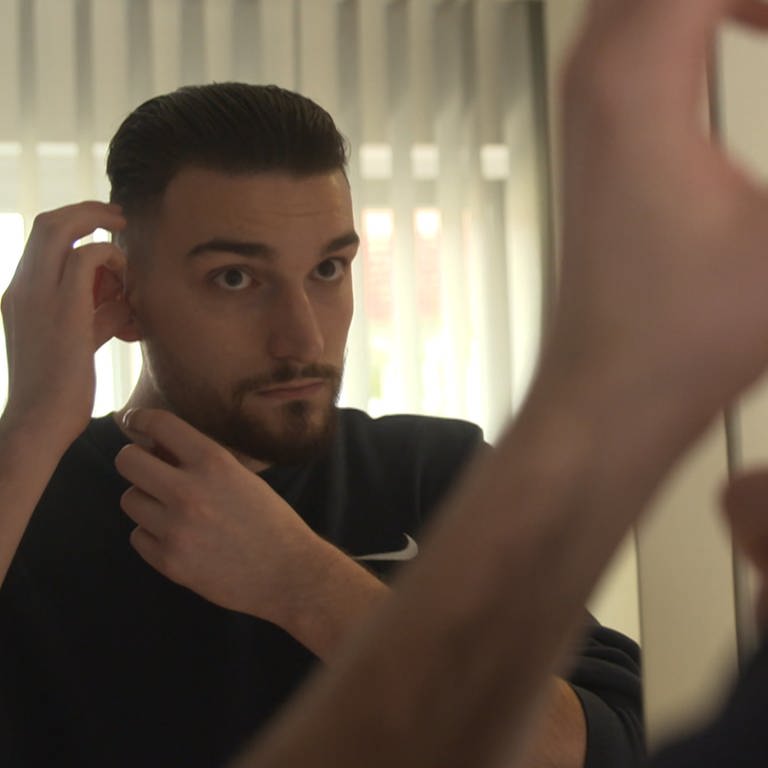 Arianit, ein junger Mann mit Vollbart, steht vor dem Spiegel und setzt sich ein Hörgerät ein (Foto: SWR)