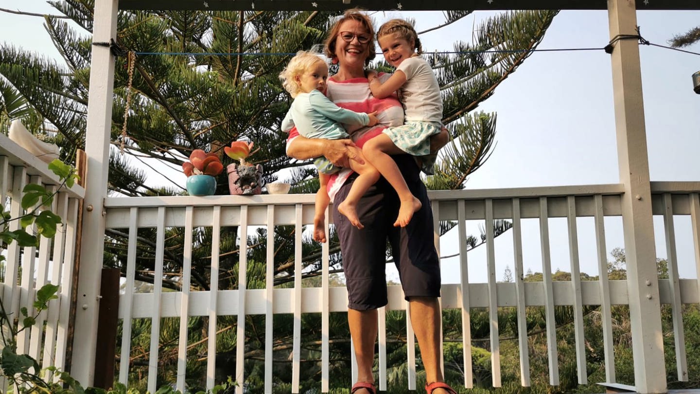 Ältere Dame mit zwei Kleinkindern auf dem Arm auf einer Terrasse.