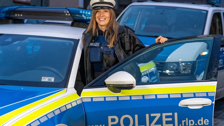 Eine junge Polizistin steht hinter der offenen Tür ihres Dienstwagens und lächelt
